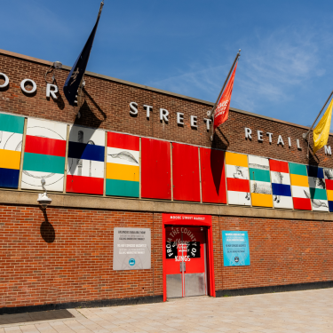 Exterior of Moore Street Retail Market, a brick building with colorful geometric panels and various flags. The entrance is marked by a red frame, flanked by informational signs. The sky is clear and blue.