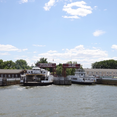 Two ferries docked at Governors Island port with surrounding buildings, trees, and a clear blue sky.