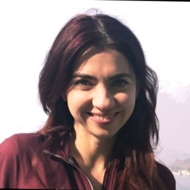Georgiana Dragu with long dark hair, wearing a maroon zip-up jacket and smiling at the camera. The background is bright and slightly blurred.