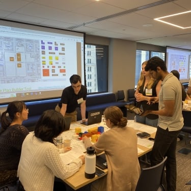 A group of NYCEDC interns dive into a collaboration workshop with the Urban Land Institute in a conference room. Some are seated around a table with papers, while others stand and discuss. A large screen displays a site plan builder tool in the background. A group of NYCEDC interns dive into a collaboration workshop with the Urban Land Institute in a conference room. Some are seated around a table with papers, while others stand and discuss. A large screen displays a site plan builder tool in the background.