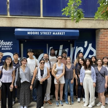 A diverse group of NYCEDC interns stand smiling in front of Moore Street Market. The storefront features blue and white signage. The group is casually dressed, and the background shows a brick wall and part of a colorful mural. A diverse group of NYCEDC interns stand smiling in front of Moore Street Market. The storefront features blue and white signage. The group is casually dressed, and the background shows a brick wall and part of a colorful mural.