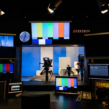 A television production control room with several monitors displaying test patterns and live feeds. A digital clock shows 23:59:58. Two cameras on tripods are in a studio setting visible through a window. A television production control room with several monitors displaying test patterns and live feeds. A digital clock shows 23:59:58. Two cameras on tripods are in a studio setting visible through a window.