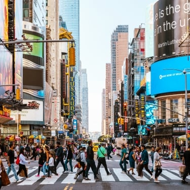 A bustling Times Square street scene in New York City with numerous people crossing at a crosswalk. Neon advertisements and tall buildings line the streets. The sky is clear, and the atmosphere is lively and vibrant.