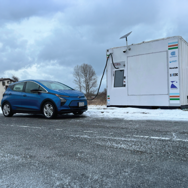 A blue electric car is parked on a snowy roadside next to a white charging station. The sky is overcast, and leafless trees are visible in the background. The charging station features logos and a cable plugged into the car. A blue electric car is parked on a snowy roadside next to a white charging station. The sky is overcast, and leafless trees are visible in the background. The charging station features logos and a cable plugged into the car.