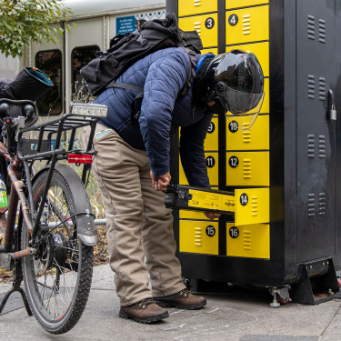 A person wearing a helmet, backpack, and blue jacket bends over to access a bright yellow charging station with numbered compartments to charge a lithium-ion battery at a pilot location in Cooper Square, Manhattan. A bicycle with a rear rack is parked nearby on a city street. A person wearing a helmet, backpack, and blue jacket bends over to access a bright yellow charging station with numbered compartments to charge a lithium-ion battery at a pilot location in Cooper Square, Manhattan. A bicycle with a rear rack is parked nearby on a city street.
