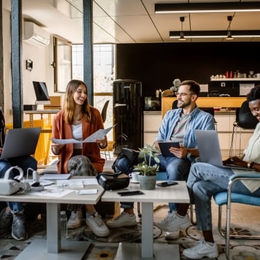 Four people sitting in a modern office, engaged in a casual meeting. They are using laptops and holding papers. The setting includes a coffee table, plants, and a kitchen area in the background, creating a relaxed atmosphere.