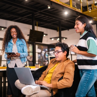 Three people in a modern office: one seated man with a laptop, a woman standing beside him with a coffee cup, and another woman smiling and holding a tablet. They appear engaged in a casual discussion.