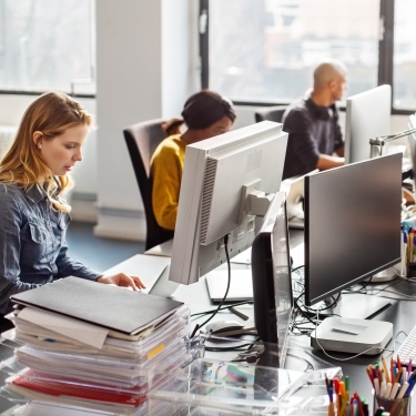 A group of people are working at desks with computers in a bright, modern office space. Large windows provide natural light. There are stacks of files and office supplies on the desks.