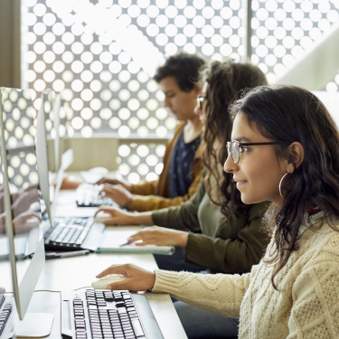 Three people working at computers in a modern office with large windows. The setting is bright due to natural light. The focus is on a person in a sweater and glasses in the foreground, with others concentrated on their screens in the background. Three people working at computers in a modern office with large windows. The setting is bright due to natural light. The focus is on a person in a sweater and glasses in the foreground, with others concentrated on their screens in the background.