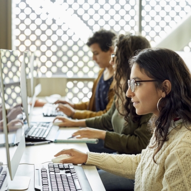 Three people working at computers in a modern office with large windows. The setting is bright due to natural light. The focus is on a person in a sweater and glasses in the foreground, with others concentrated on their screens in the background.