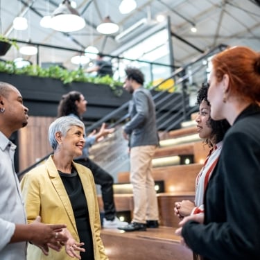 A group of diverse people engage in conversation on wooden bleachers in a modern office space. Green plants adorn the upper levels. Some people are standing while others sit or ascend the staircase in the background.