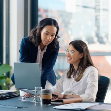 Two women in an office setting. One is standing and pointing at a laptop screen, while the other is seated, smiling and looking at the screen. There are notebooks and a coffee cup on the table. Large windows are in the background.