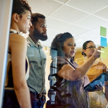 A group of four startup employees stand in an office, discussing and placing colorful sticky notes on a glass wall. One person is pointing while holding a tablet.