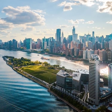 Aerial view of the New York City skyline with tall buildings reflecting sunlight. A river flows alongside a green park on the left, with a curved shoreline and modern structures nearby. The sky is partly cloudy, creating a serene, bright atmosphere.