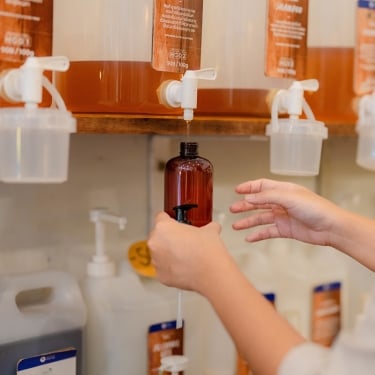 A person is refilling a brown bottle from a wall-mounted dispenser at a bulk store. Several dispensers with different liquids are visible, each with white taps. The person holds the bottle upright, focusing on the refill process.