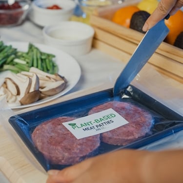 A person opens a package of plant-based meat patties with a knife. The table is set with various fresh ingredients, including mushrooms, green beans, sliced tomatoes, avocadoes, and grapes.