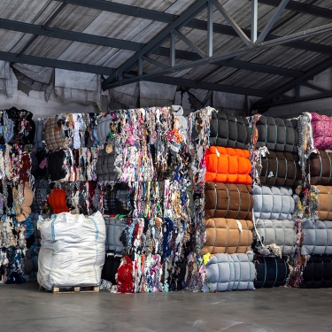 Stacks of large bales of mixed, colorful textiles and fabrics are stored in a spacious industrial warehouse. They are neatly arranged against a white brick wall, under a metal roof structure.