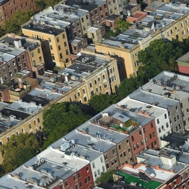 Aerial view of several city blocks with rows of apartment buildings featuring white roofs, separated by tree-lined streets in an urban neighborhood.