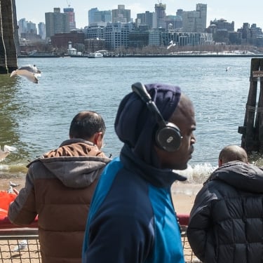 Three men stand near a waterfront railing, with one in the foreground wearing headphones. Seagulls fly nearby, and a city skyline is visible across the water under a partly cloudy sky.