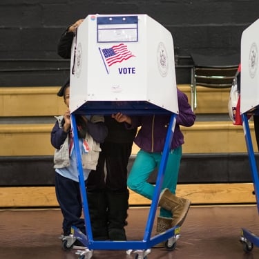Three voting booths with “VOTE” and an American flag are occupied by people casting ballots in a gymnasium. Some children are visible, including one peeking from under a booth. Wooden bleachers are in the background.