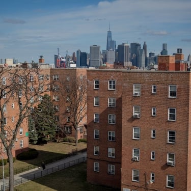 Red brick apartment buildings with small lawns and leafless trees in the foreground; the New York City skyline with skyscrapers and the One World Trade Center is visible in the background under a partly cloudy sky.