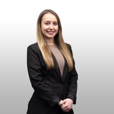 Woman with long, straight blonde hair wearing a black blazer over a beige top stands smiling with her hands clasped in front of her against a light gray, gradient background. Woman with long, straight blonde hair wearing a black blazer over a beige top stands smiling with her hands clasped in front of her against a light gray, gradient background.