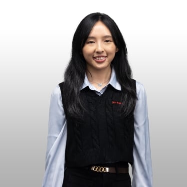 Young woman with long black hair smiles at the camera. She is wearing a light blue blouse under a black sweater vest and black pants with a gold-buckled belt, standing against a white-to-gray gradient background. Young woman with long black hair smiles at the camera. She is wearing a light blue blouse under a black sweater vest and black pants with a gold-buckled belt, standing against a white-to-gray gradient background.