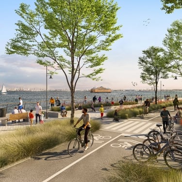 People walk, cycle, and relax along a busy waterfront path lined with trees and benches. A ferry and sailboat are visible on the water, and the city skyline can be seen in the distance under a blue sky. People walk, cycle, and relax along a busy waterfront path lined with trees and benches. A ferry and sailboat are visible on the water, and the city skyline can be seen in the distance under a blue sky.