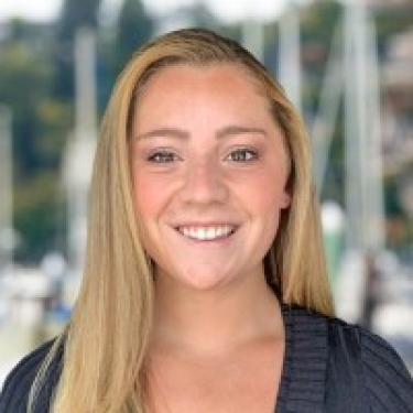 Brynna O’Leary with long blonde hair in a dark top, smiling and standing in front of a blurred marina backdrop with sailboats.