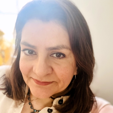 Claudia Franco with shoulder-length brown hair wearing a cream-colored blouse and a beaded necklace, smiling softly at the camera in a relaxed setting.