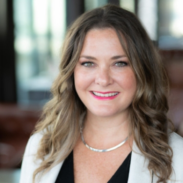 Kristy Sisko Wallace with wavy brown hair, smiles warmly at the camera. She is wearing a light-colored blazer over a black top and a silver necklace. The background is softly blurred, suggesting an indoor setting.