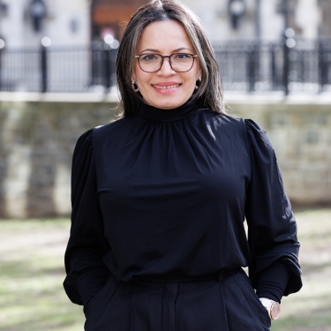 Nury M. Gutierrez smiling in front of a historic building. She is wearing glasses and navy blouse and pants, with her hands in her pockets. The background features stone architecture and a grassy area.