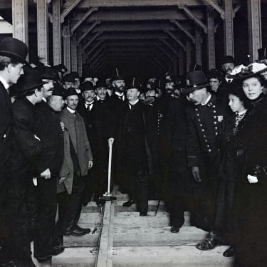 A black-and-white photo shows a crowd of men and women, some in hats and uniforms, gathered around a subway track inside a tunnel during a ceremonial event. Mayor Seth Low (center) hammers in spike for IRT subway, ca. 1902-04.