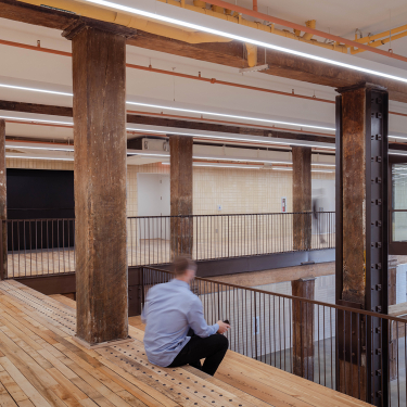 A person sits on a wooden bench in an industrial-style interior with exposed beams, wooden floors, metal railings, and cream tile walls, creating a spacious, modern atmosphere.