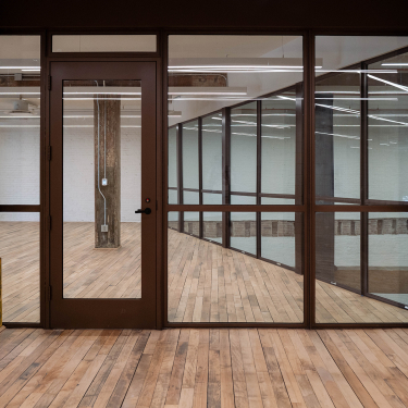 A modern, empty office space with floor-to-ceiling glass walls, a glass door, exposed concrete pillars, and light wood flooring. A sign outside the office displays the number 200. The room is well-lit with ceiling lights.