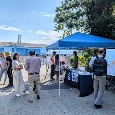 People gather at Brooklyn Bridge Park near a blue tent with an NYC/EDC table, engaging in conversation and viewing maps and posters under sunny skies, surrounded by greenery and industrial buildings. People gather at Brooklyn Bridge Park near a blue tent with an NYC/EDC table, engaging in conversation and viewing maps and posters under sunny skies, surrounded by greenery and industrial buildings.