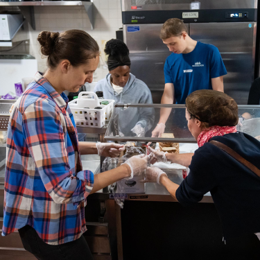Six people stand behind a counter serving food in a cafeteria or soup kitchen, wearing gloves and focused on preparing and distributing meals. Various trays of food are visible in front of them.