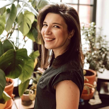Joanna Kaniewska with straight brown hair, smiles while standing indoors next to large leafy plants and potted greenery, with natural light coming through a window in the background.