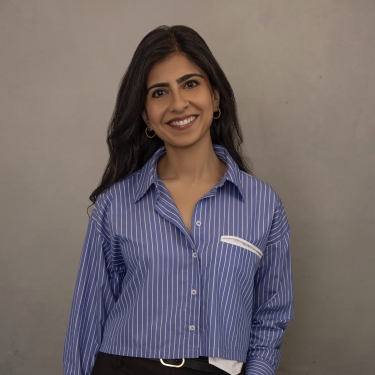 Shefali Lakhani with long dark hair smiles at the camera. She is wearing a blue and white striped button-up shirt and dark pants, standing against a plain, light gray background.