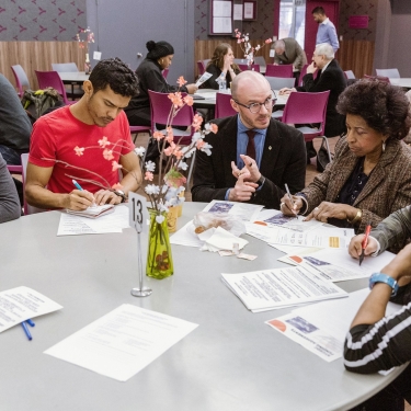 A group of diverse people at the Non-Profit Coffee hour sit around a round table in a community setting, engaged in discussion. Papers, pens, and a vase with flowers are on the table. Other people are visible in the background, seated at additional tables. Photo by Kreg Holt/NYCEDC. A group of diverse people at the Non-Profit Coffee hour sit around a round table in a community setting, engaged in discussion. Papers, pens, and a vase with flowers are on the table. Other people are visible in the background, seated at additional tables. Photo by Kreg Holt/NYCEDC.
