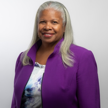 Deneen Donnley with long gray hair, wearing a purple blazer over a patterned top, smiling against a plain white background.