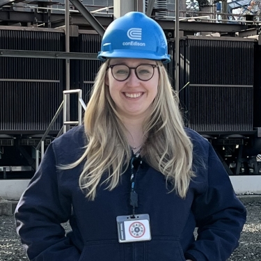 Kayla Mallay smiling outdoors and wearing glasses, a blue Con Edison hard hat, and a navy jacket. She has long blonde hair and a lanyard with an ID badge. Industrial equipment and structures are in the background.