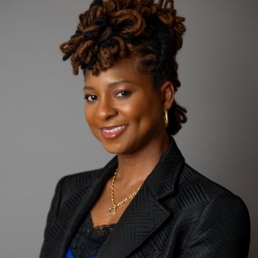 Nicole Mosby with styled locs, wearing a black blazer over a blue top, gold necklace, and hoop earrings, smiles at the camera against a plain gray background.