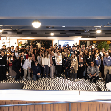 NYC Startup Internship program participants pose for a group photo indoors, many wearing name tags and business attire, standing on a black-and-white patterned floor in a spacious, modern room.