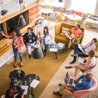 A woman stands at a blackboard leading a discussion with a group of people seated in a modern, brightly lit meeting space. Attendees are taking notes, using laptops, and listening attentively.