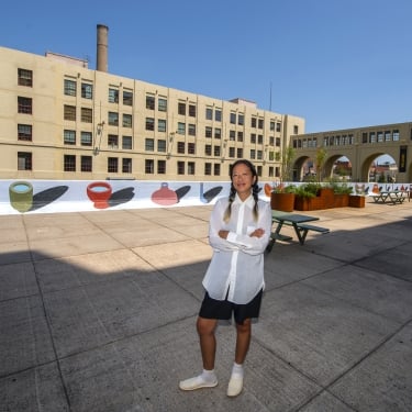 Brooklyn artist Crys Yin stands with arms crossed on a sunlit plaza, featuring colorful her mural Full Form on low walls, outdoor seating, and a large industrial-style building in the background under a clear blue sky.