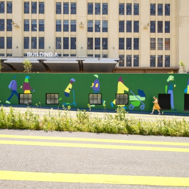 A green mural on a wall depicts colorful silhouettes of people walking and pushing strollers. A person in an orange outfit walks past in front of the mural. Behind the wall is a beige industrial building labeled “Building A.”.