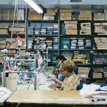 Three women work at a table in a workshop filled with shelves of organized boxes and craft supplies. The workspace is busy, with tools and materials spread out in front of them.