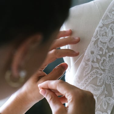 A person sews delicate lace fabric onto a white dress on a mannequin, focusing intently on their work and using both hands to guide the needle and fabric.