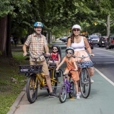 A family of four wearing helmets stands with their bicycles on a bike path surrounded by trees. The parents have adult bikes, one child rides a small bike, and the other sits in a child seat. They are all smiling.